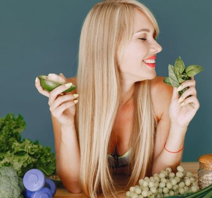 A smiling woman holding avocado and basil, surrounded by fresh vegetables and fruits for healthy eating.